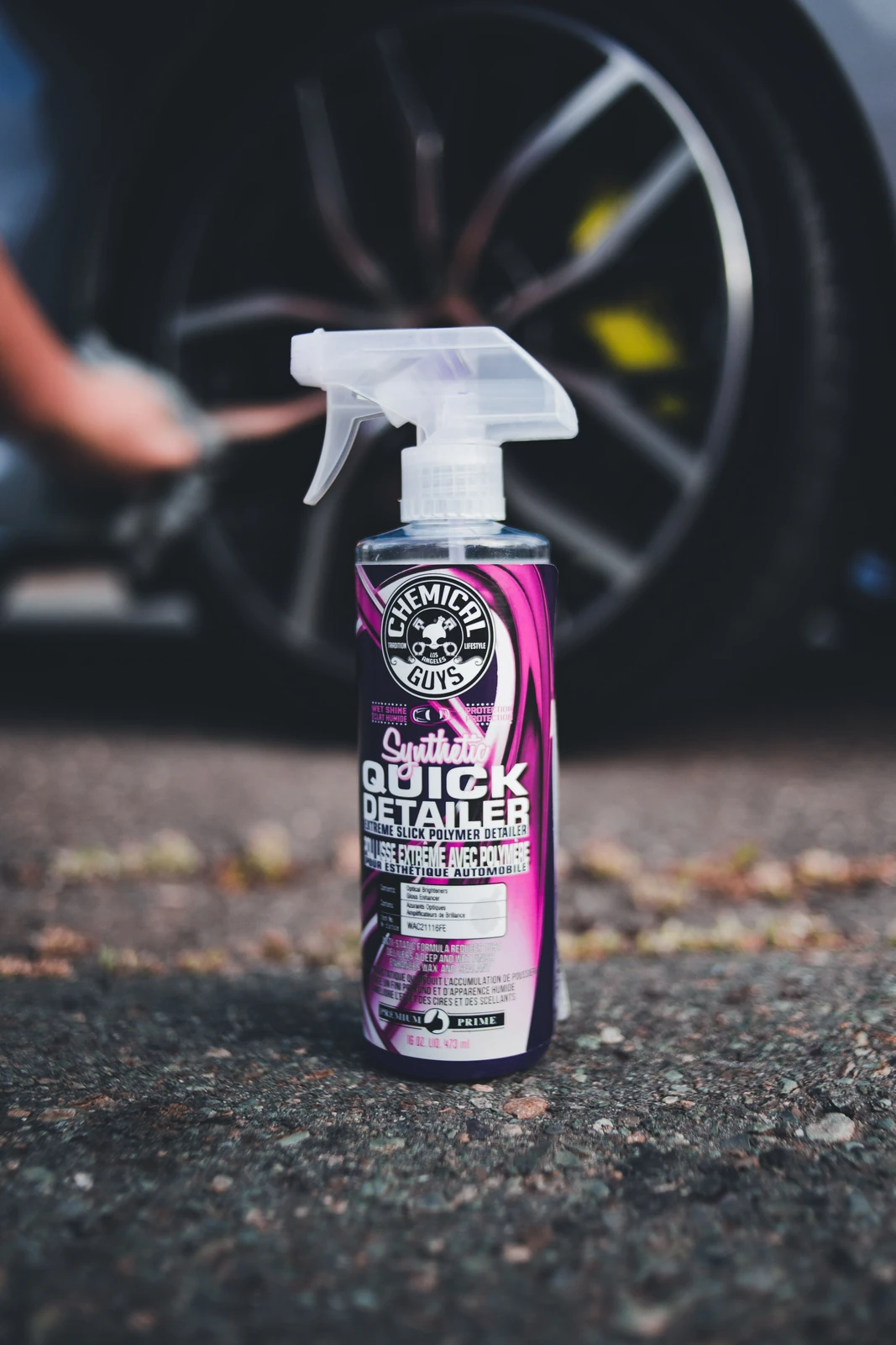 Car-care detailing spray bottle in front of a softly focused vehicle wheel, illustrating automotive detailing products; stock imagery, not a Meitar product or operation.