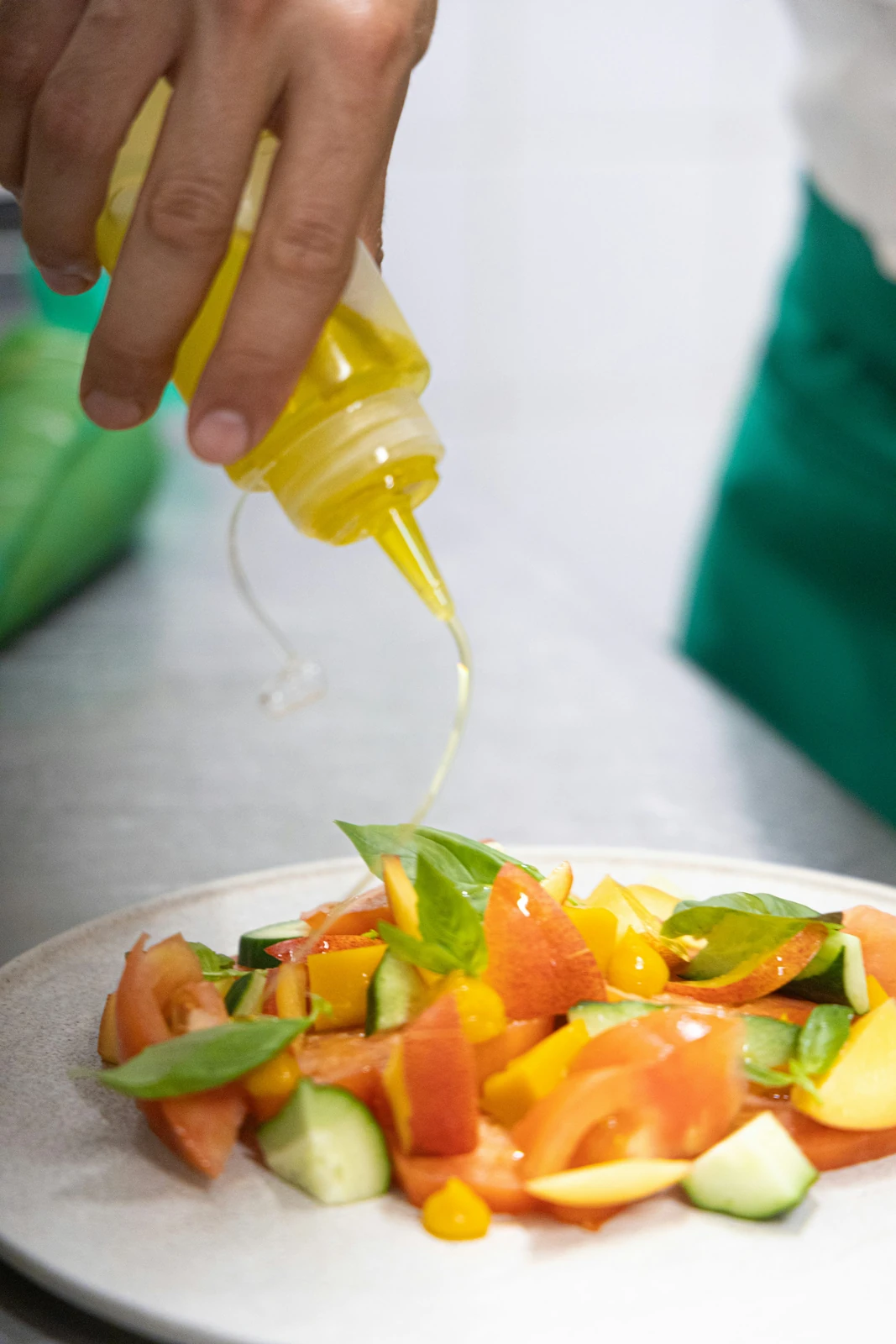 A hand pours golden cooking oil from a bottle over a plate of fresh chopped vegetables and leafy greens.