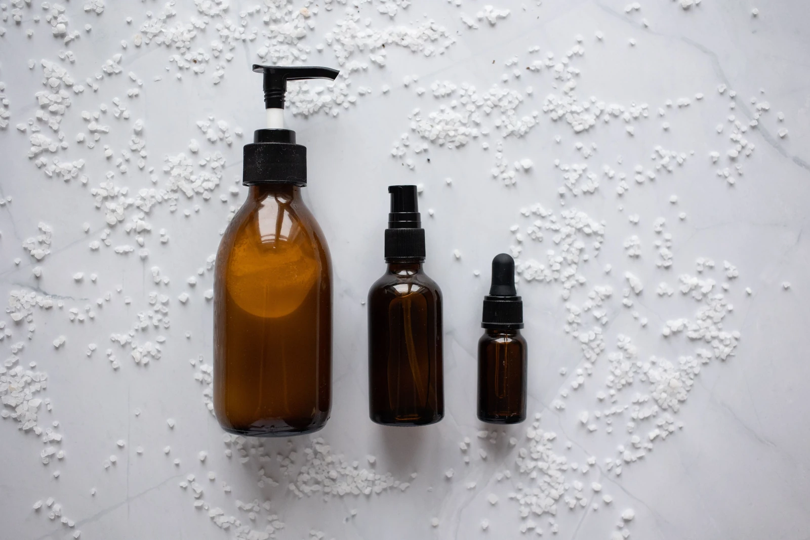 Brown glass skincare bottles with pumps and droppers arranged on a light marble surface with scattered salt crystals — illustrative personal care and cosmetics product imagery, not a Meitar product.